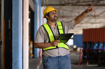 A seriously bearded warehouse manager wearing a yellow hard hat and green vest carries a clipboard and counts boxes while inspecting merchandise.