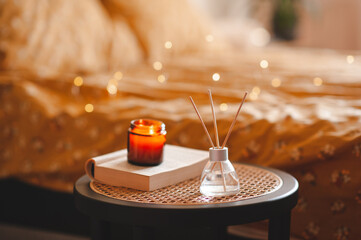 Home liquid perfume in glass bottle with bamboo sticks and burning candle on paper book staying on rattan table close up in bedroom. Cozy atmosphere. - obrazy, fototapety, plakaty