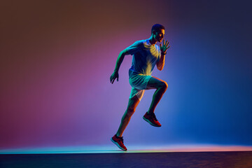 Young athletic man with muscular body, in headphones, training, running on gradient pink blue background in neon light. Concept of active and healthy lifestyle, sport, hobby, motivation, endurance
