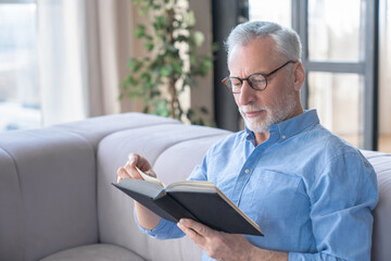 Close up portrait of an elderly old mature senior handsome man reading a book sitting a couch sofa...