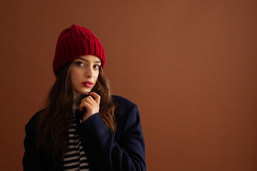 Fashionable confident woman wearing stylish red knitted beanie hat, sweater, classic blue woolen coat, posing on brown background. Studio fashion portrait. Copy, empty, blank space for text  - 791768971