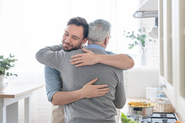Cute loving caring adult caucasian son embracing hugging his old elderly senior father in the kitchen while cooking lunch, dinner, preparing meal together. Happy father`s day! I love you, dad! - obrazy, fototapety, plakaty