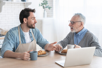 Communicating talking old elderly senior father with his adult son having breakfast using laptop in...