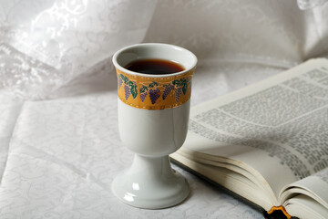 Porcelain goblet filled with wine resting on an open book. The goblet is adorned with a decorative band featuring grape illustrations. The scene is set against elegant, white lace curtains tablecloth