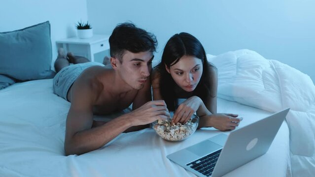 A couple relaxes in bed while watching a movie on a laptop and enjoying a bowl of popcorn at night.