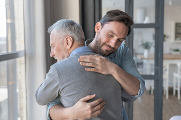 Caring cute caucasian adult son hugging embracing his old elderly senior father showing him love...