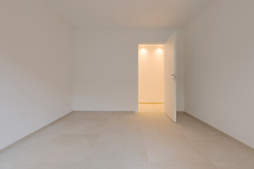 Inside an empty room and to the right a door leading to the corridor with a light on. All the walls and ceiling are white. - 791804145