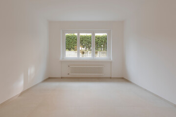Empty interior of a room, with a central window in the background. Under the window a radiator.