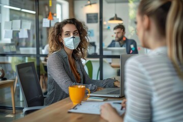 A woman wearing a protective mask sits at a table in an office, engaging in a discussion with an unseen colleague