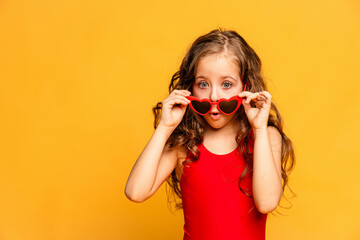 Little child girl in red swimsuit and red funny summer sunglasses surprised expression posing on...