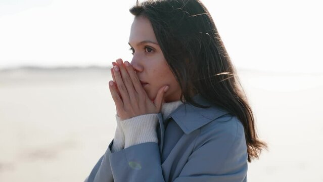 Young woman in a coat warms her hands, gazing at the sea on a chilly day, embodying solitude and reflection at the beach.