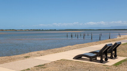 Two public wooden lounge chairs on a concrete pathway overlooking a peaceful tidal inlet beach at...