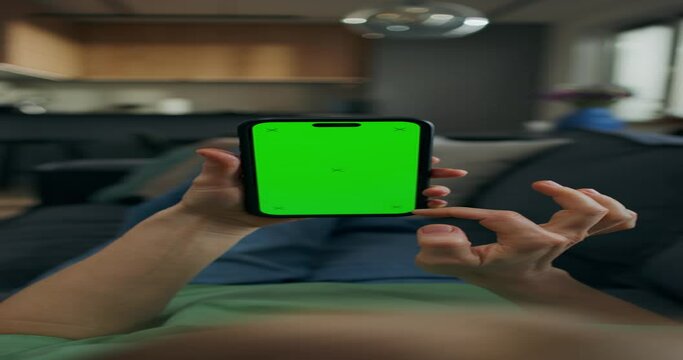 A woman uses a mobile phone with green screen lying on the couch at home. A rear view of her hands, an unrecognizable person