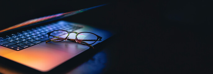 Glasses lie on the laptop, reflecting light from the screen in the dark. - obrazy, fototapety, plakaty