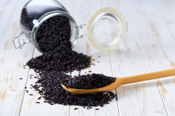 Raw black rice in wooden scoop and in glass storage jar on a white wooden table.