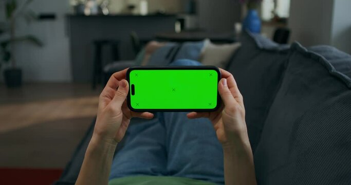 A woman holds a mobile phone with green screen in her hand. She is lying on the couch at home, close-up of her hands, unrecognizable person