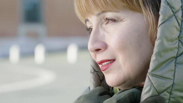 Wearing a hood over her head, a middle-aged woman with red hair smiles melancholically in the public square, her smile bittersweet against the backdrop of everyday life