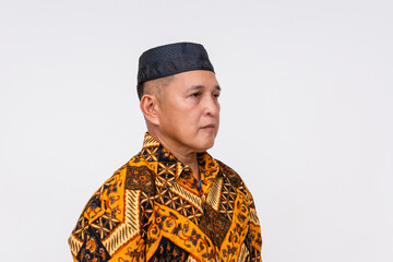 Portrait of a sober Indonesian man wearing a traditional batik shirt and a songkok, against a white...