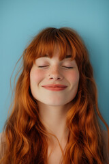 A young woman with long, flowing red hair closes her eyes in a moment of bliss, a slight smile on her lips, against a soft blue background.