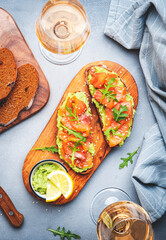 Avocado sandwich with salmon on rye bread with guacamole sauce, arugula and sesame seeds on wooden board, rose wine glass on gray table background, top view - 791862327