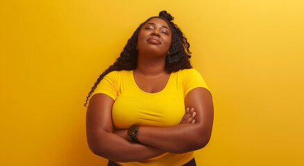 Obraz premium A confident plus-size woman in a bright yellow shirt stands with her arms crossed, eyes closed, and a serene smile on her face against a yellow backdrop.
