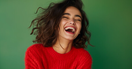 A woman in a textured red sweater laughs with abandon, her brunette hair dancing in the air, epitomizing infectious joy against a green canvas.