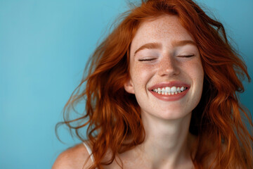 Close-up of a freckled redhead woman with flowing hair and a radiant smile, her eyes shut in joy against a refreshing blue background.