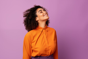 A woman with a playful smile looks up, her mood uplifted, dressed in a lively orange shirt which beautifully contrasts with the lilac background.