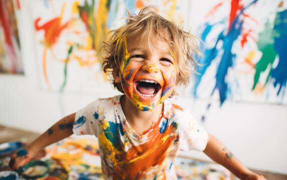 A young child is the epitome of joy and creativity, his face and clothes a canvas of vibrant paint splatters, against a backdrop of colorful artwork.