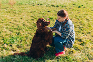 Cheerful happy young woman playing with her dog in the yard in summer. Beautiful Irish Setter dog is lying in grass. Pets adoption, care and love concept