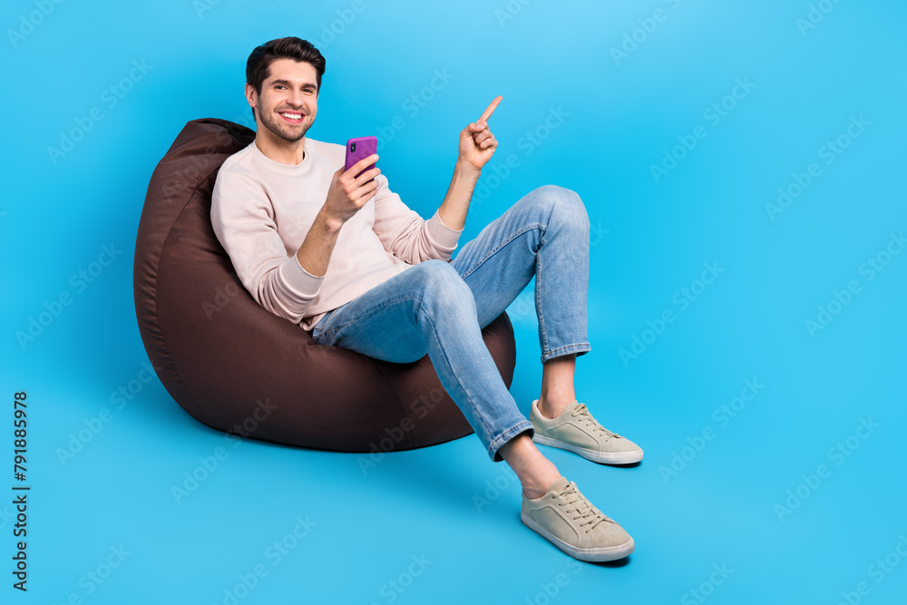 Poster full length photo of nice guy wear pullover sit on pouf indicating at eshop empty space hold smartph