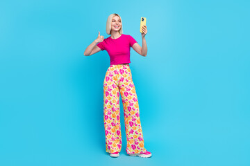 Full body photo of attractive young woman hold telephone selfie thumb up dressed stylish pink clothes isolated on blue color background