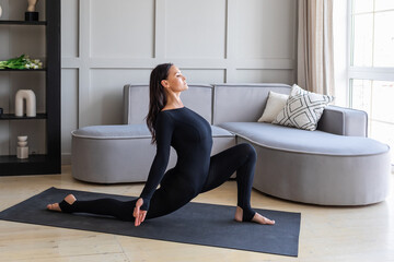 Woman practicing yoga in the room, performing a variation of Anjenyasana exercise, half moon pose, working out in black sportswear on a mat, full length view - obrazy, fototapety, plakaty