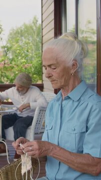 Medium vertical shot of pensive old Caucasian woman sitting in armchair on terrace at retirement facility, on summer day, knitting, looking away, male residents playing chess in backgroundMedium verti