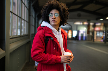 Curly-haired young guy at the railway platform with a coffee cup in hands