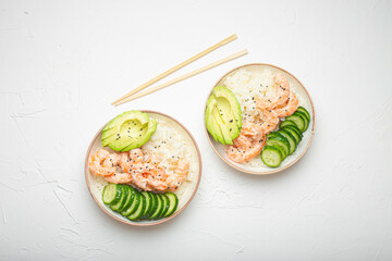 Two white ceramic bowls with rice, shrimps, avocado, vegetables and sesame seeds and wooden sticks on white concrete stone background top view. Healthy asian style poke bowl.