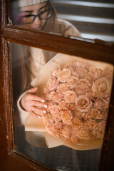 Girl in sweater stands behind glass in a wooden frame with a beautiful roses - 791928783