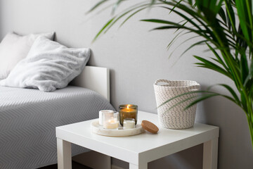Home decor, stylish interior. Burning candles on marble tray standing on white badside table in bedroom.  Bed with grey blanket and pillows, wicker basket, howea palm plant.