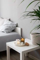 Home decor, stylish interior. Burning candles on marble tray standing on white badside table in bedroom.  Bed with grey blanket and pillows, wicker basket, howea palm plant.