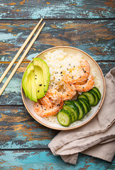 White ceramic bowl with rice, shrimps, avocado, vegetables and sesame seeds and chopsticks on colourful rustic wooden background top view. Healthy asian style poke bowl.