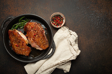 Two roasted duck breast fillets with crispy skin, with pepper and rosemary, top view in black cast iron pan with knife, dark brown concrete rustic background, space for text.