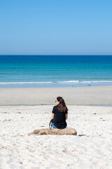 A serene image capturing a lone woman sitting on a log at the beach, gazing out towards the endless blue ocean under a clear sky