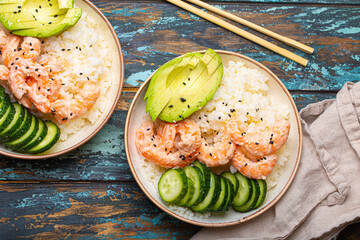 Two white ceramic bowls with rice, shrimps, avocado, vegetables and sesame seeds and chopsticks on colourful rustic wooden background top view. Healthy asian style poke bowl.
