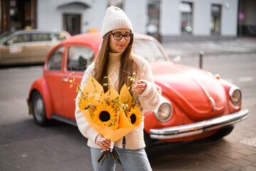 Retro car and a young girl in glasses and sweater stands with a bouquet - 791932730