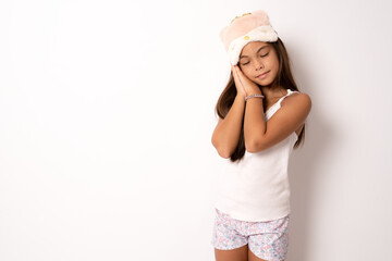 Beautiful child girl wearing sleep mask and pajama standing over isolated white background sleeping...