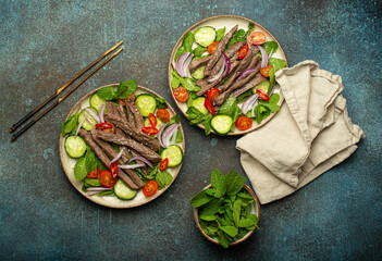 Two plates with traditional Thai beef salad with vegetables and mint top view served on rustic concrete background, healthy exotic asian meal.