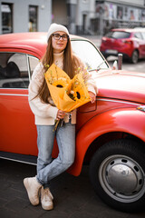 Young girl in sweater and jeans stands near a red vintage car on a street with a flowers