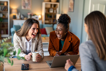 Three diverse female colleagues laughing in a modern office.