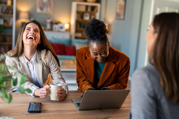 Three female colleagues laughing while having coffee and working on a laptop in a modern office.