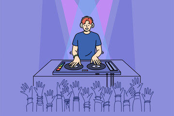 Man DJ performs in nightclub spinning records on mixing console, near crowd of partygoers with hands raised. Cheerful guy DJ in headphones plays dance hits for party or festival visitors